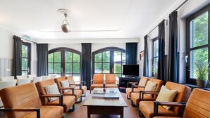 Ein geräumiges Wohnzimmer mit Ledersofas, die um einen zentralen Couchtisch angeordnet sind, großen Fenstern mit Vorhängen und einem Deckenventilator.