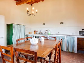 A dining area with a wooden table and chairs, a chandelier, and a kitchen in the background.