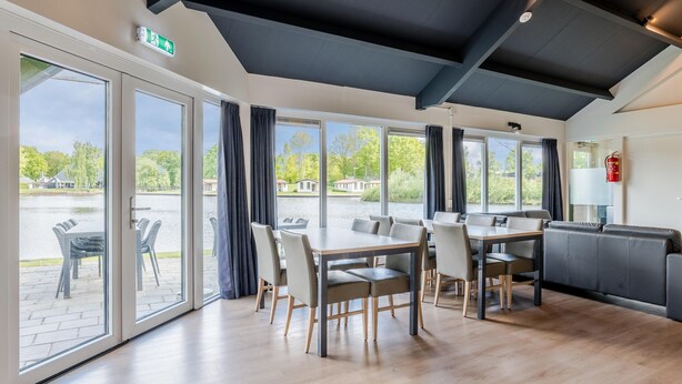 A modern dining area with a large table and chairs, large windows with curtains, and a view of a courtyard. - Well Furnished Home for Groups with Dishwasher near Water (Gasselternijveen)