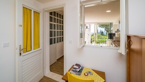Une petite pièce avec une porte en bois, un rideau jaune, une fenêtre donnant sur un balcon, et une table avec des livres et une tasse.