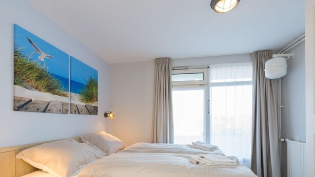 A bedroom with a large bed, a wall-mounted light, a window with curtains, and a beach-themed painting. - Apartment in Zoutelande Near Sandy Beach (Zoutelande)