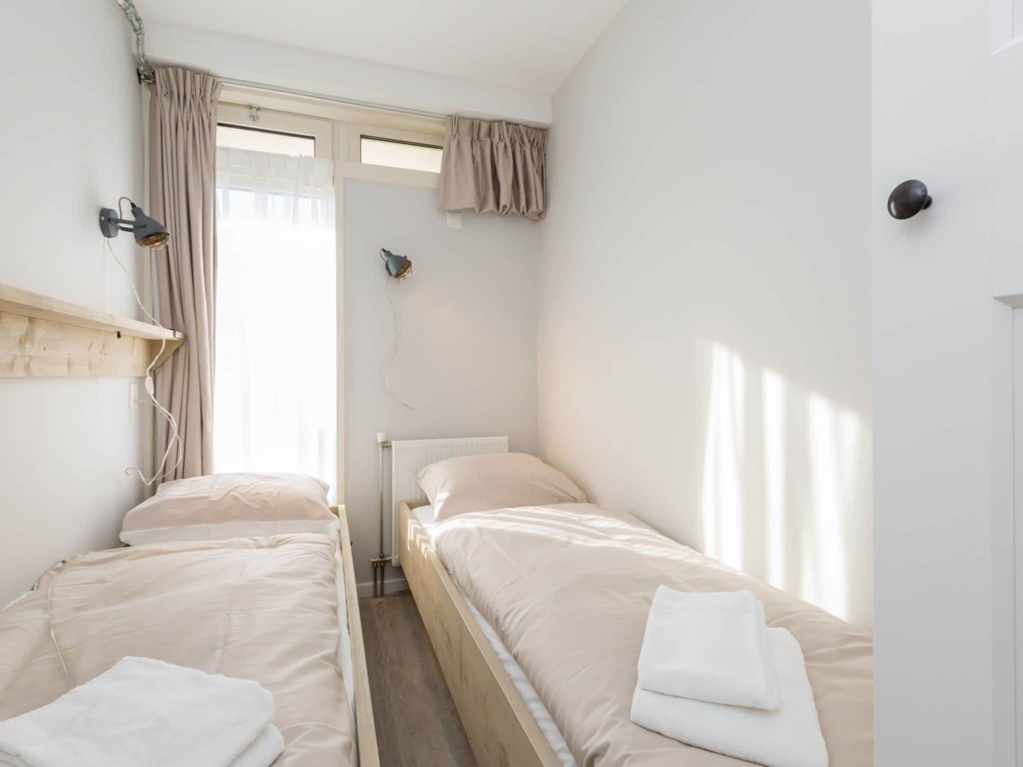 A small, clean room with two single beds, a window with curtains, a radiator, and a wall-mounted lamp.