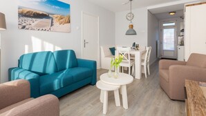 A modern living room with a teal sofa, a white dining table, and a beige armchair. A painting of a beach scene is on the wall. - Apartment in Zoutelande Near Sandy Beach (Zoutelande)