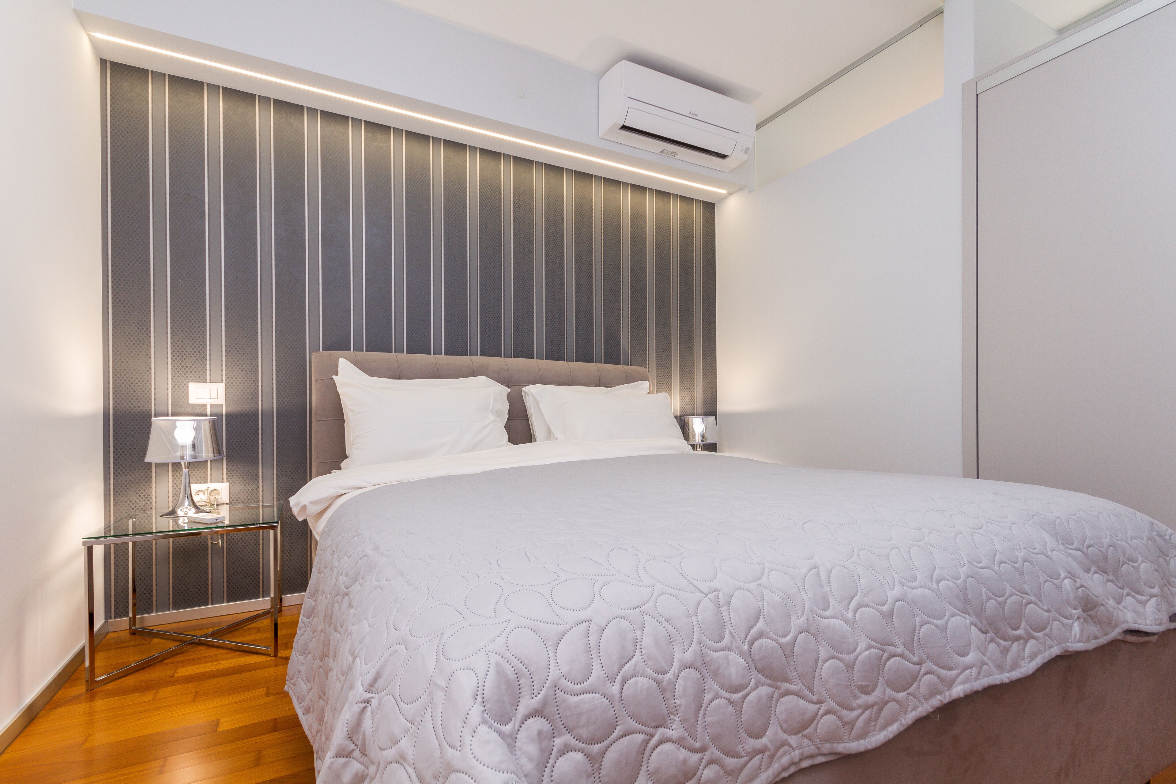 A modern bedroom with a large bed, a nightstand with a lamp, and a wall-mounted air conditioner.
