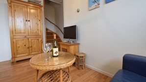 Uma sala de estar com um armário de madeira, uma mesa de vidro com taças de vinho e uma garrafa, uma televisão sobre um suporte de madeira e um sofá azul.