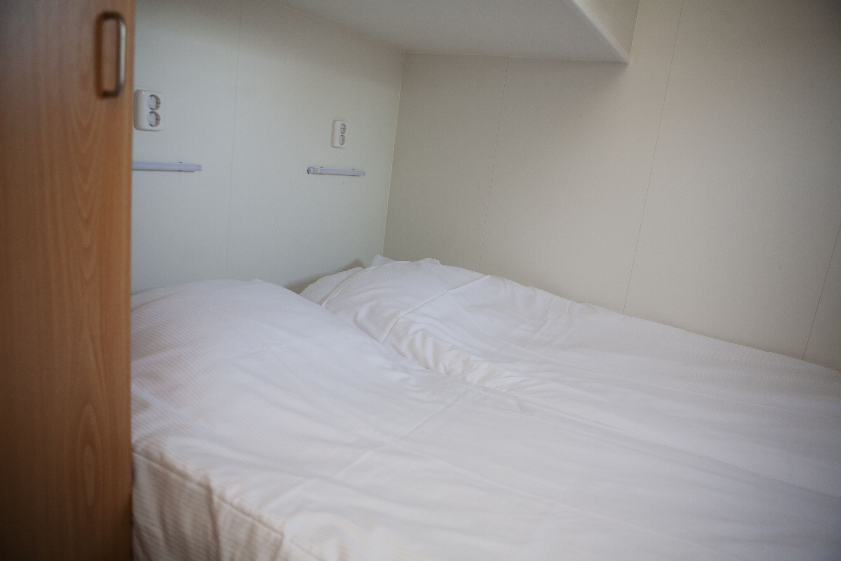 Une petite chambre avec un lit simple, une porte en bois, des murs blancs et un plafond blanc.