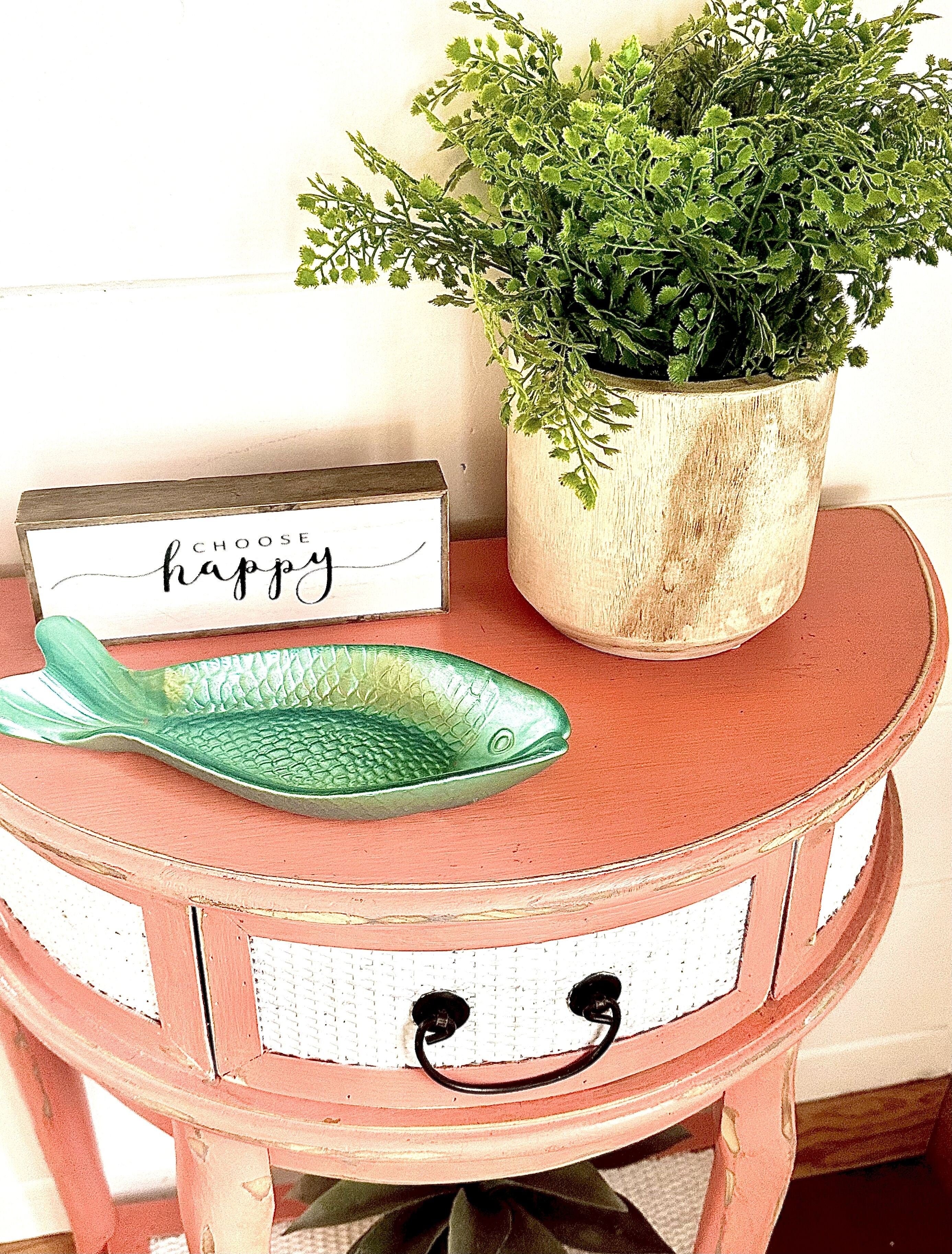 Una mesa redonda pequeña con un plato verde con forma de pez y un letrero que dice 'CHOOSE happy'.