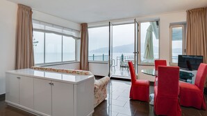 A modern hotel room with a large window offering a view of the sea, a small balcony with a table and chairs, and a TV on a stand. - Holiday Home in Meina With Private Beach (Meina)