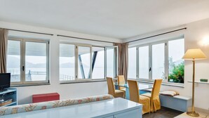 A modern hotel room with a large window, a sofa, a dining table with chairs, a TV, and a lamp. - Holiday Home in Meina With Private Beach (Meina)