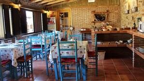 Een rustieke eethoek met houten balken, betegelde vloer en blauwe stoelen. Tafels zijn gedekt met witte tafelkleden en rode servetten.