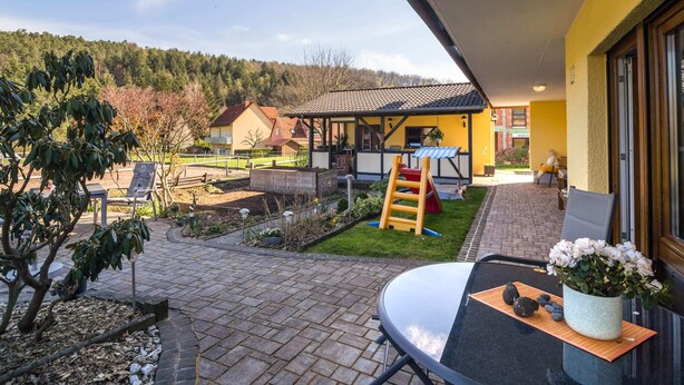 A patio area with a table, chairs, and a potted plant. There is a play area with a slide and a small house structure. - Apartment in the Odenwald With Terrace (Mossautal)