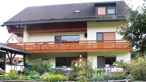 A two-story house with a balcony, surrounded by a garden with various plants and flowers. - Apartment in the Odenwald With Terrace (Mossautal)