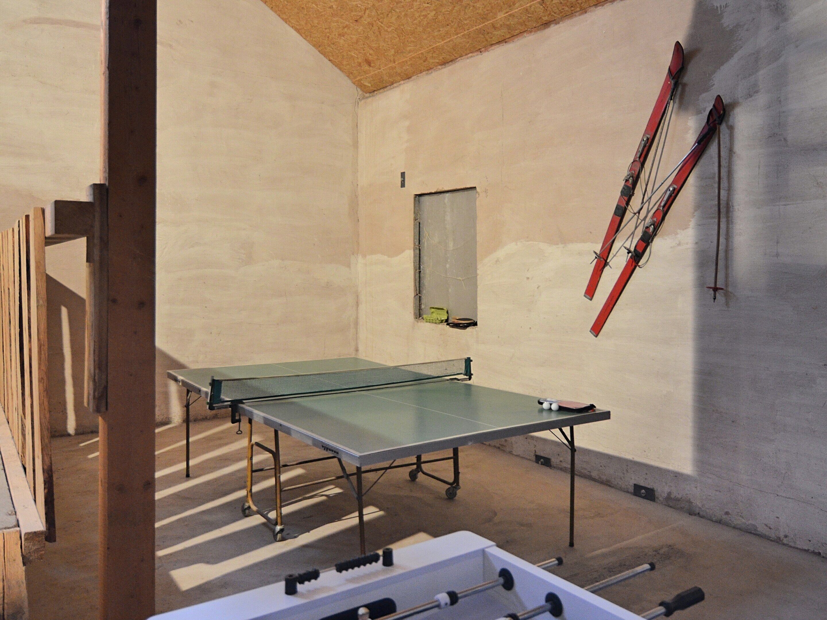 Une table de ping-pong se trouve dans une pièce dont le sol et les murs sont en béton, le plafond en bois et qui possède une fenêtre.