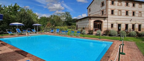A rectangular swimming pool with blue water, surrounded by a brick patio. There are blue lounge chairs and umbrellas along the poolside. A stone building with multiple windows and a door is visible in the background.