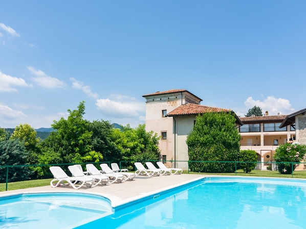 A swimming pool with white lounge chairs, a building with a tiled roof, and greenery in the background.