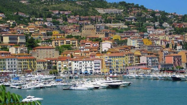 Une ville c ôtière aux bâtiments aux couleurs vives, un port de plaisance rempli de bateaux et, en arrière-plan, une colline.
