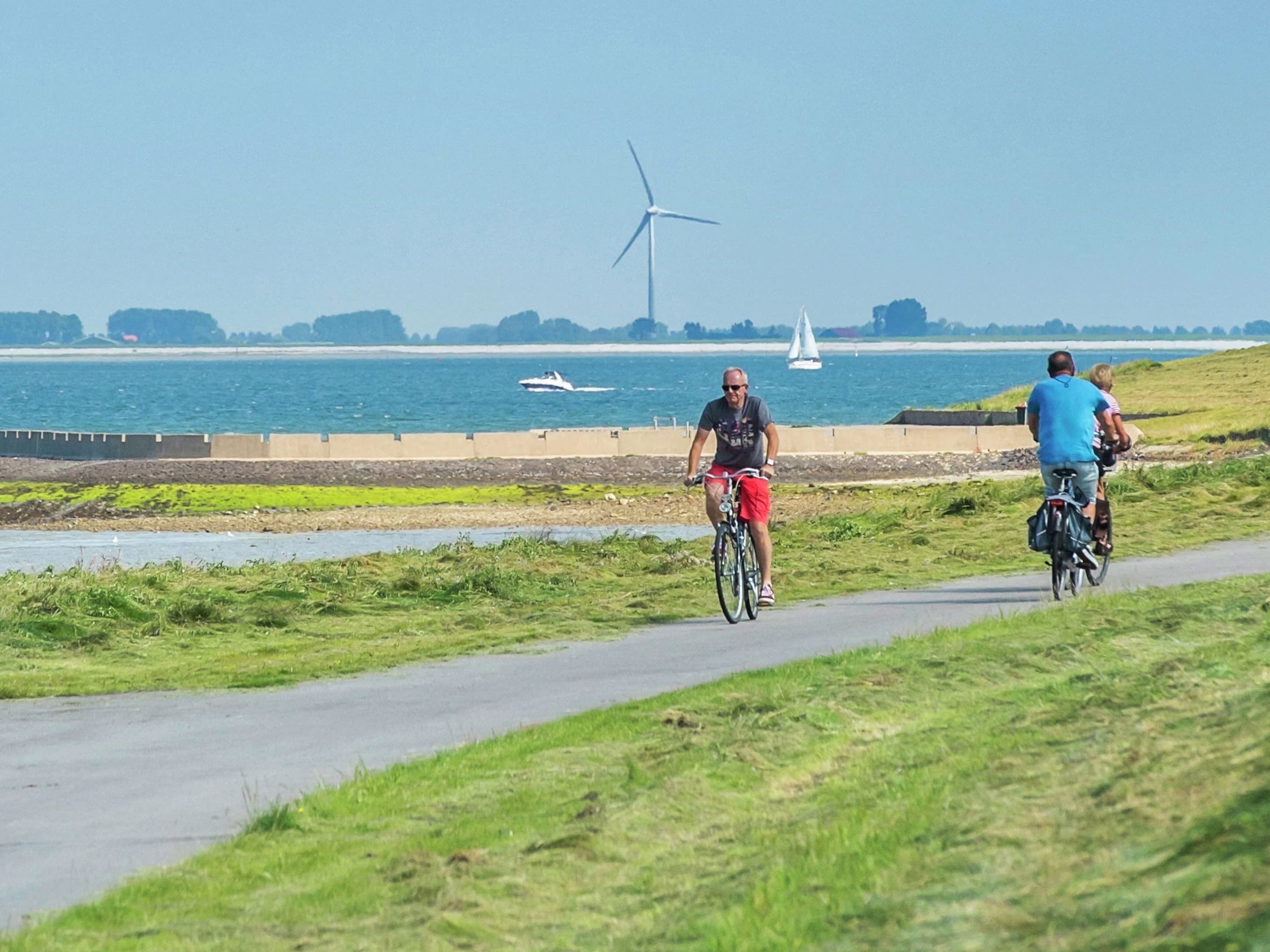 Ein befestigter Weg entlang eines Gewässers, im Hintergrund eine Windkraftanlage.