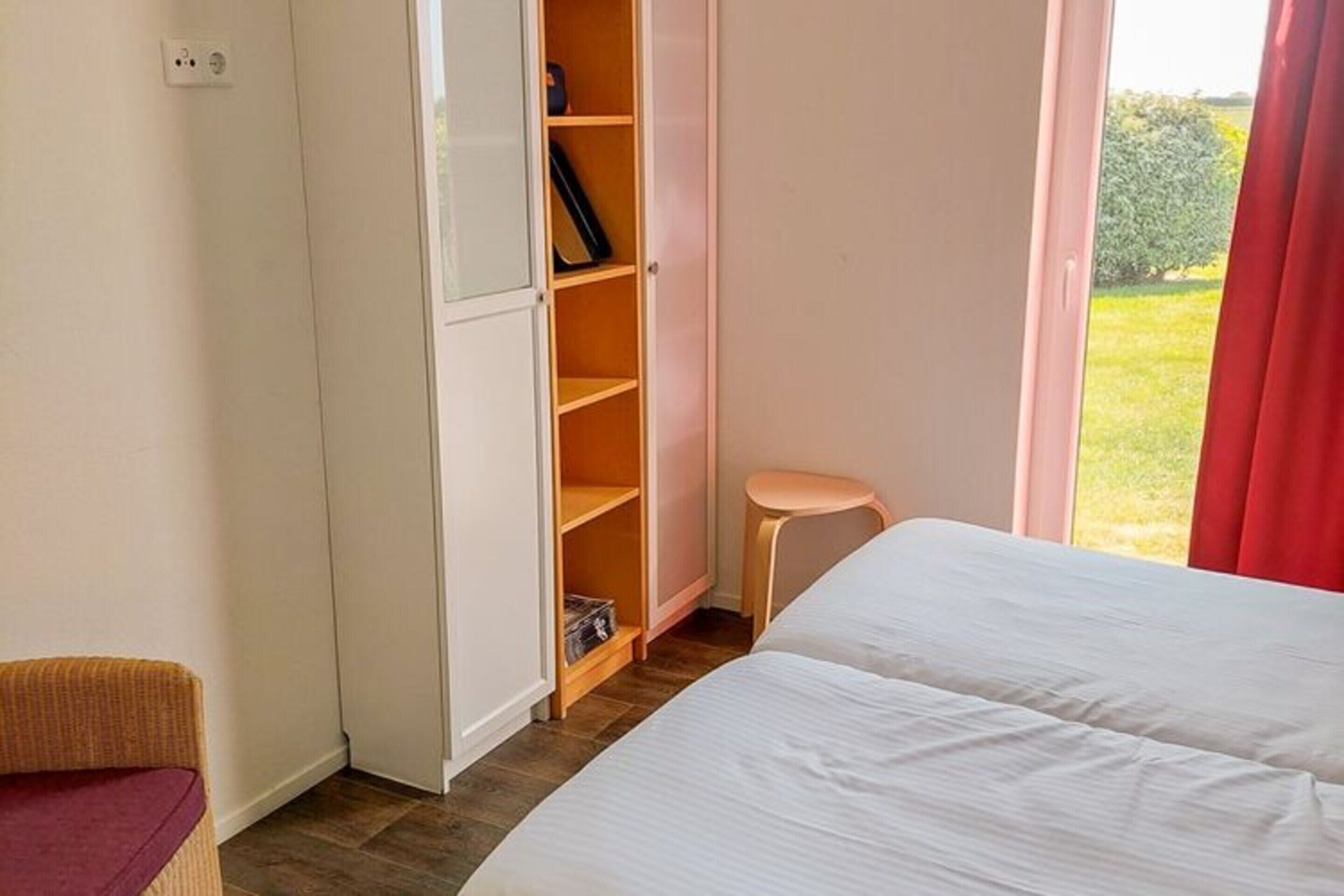 A bedroom with a bed, a wardrobe, a small stool, and a window with a view of greenery.