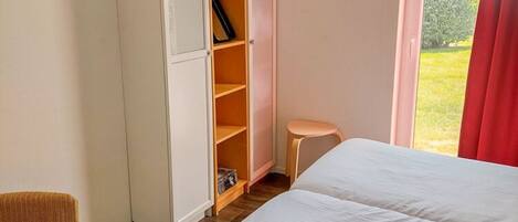 A bedroom with a bed, a wardrobe, a small stool, and a window with a view of greenery.