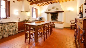 A rustic kitchen with exposed wooden beams, a central island with chairs, a fireplace, and a tiled floor.