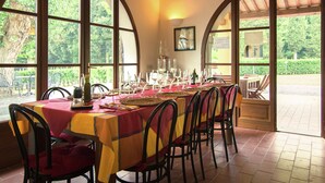 A dining area with a long table set for a meal, large arched windows, and a view of greenery outside.