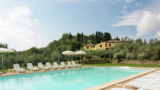 A swimming pool with white lounge chairs and umbrellas, surrounded by greenery and a hillside villa.