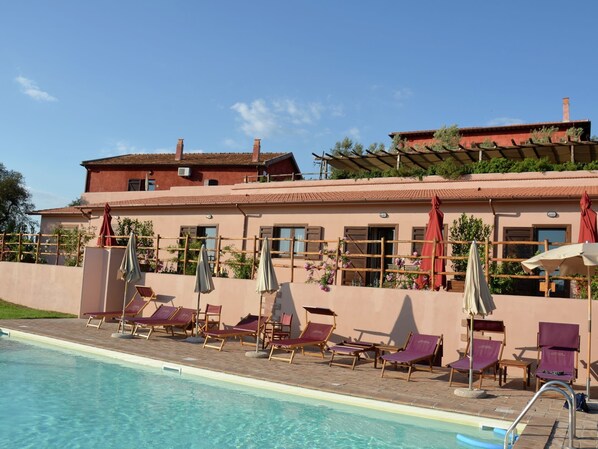 Un espace aménagé au bord de la piscine, avec des chaises longues et des parasols, une piscine à l’eau claire et un bâtiment doté d’un toit rouge et de murs de couleur beige.