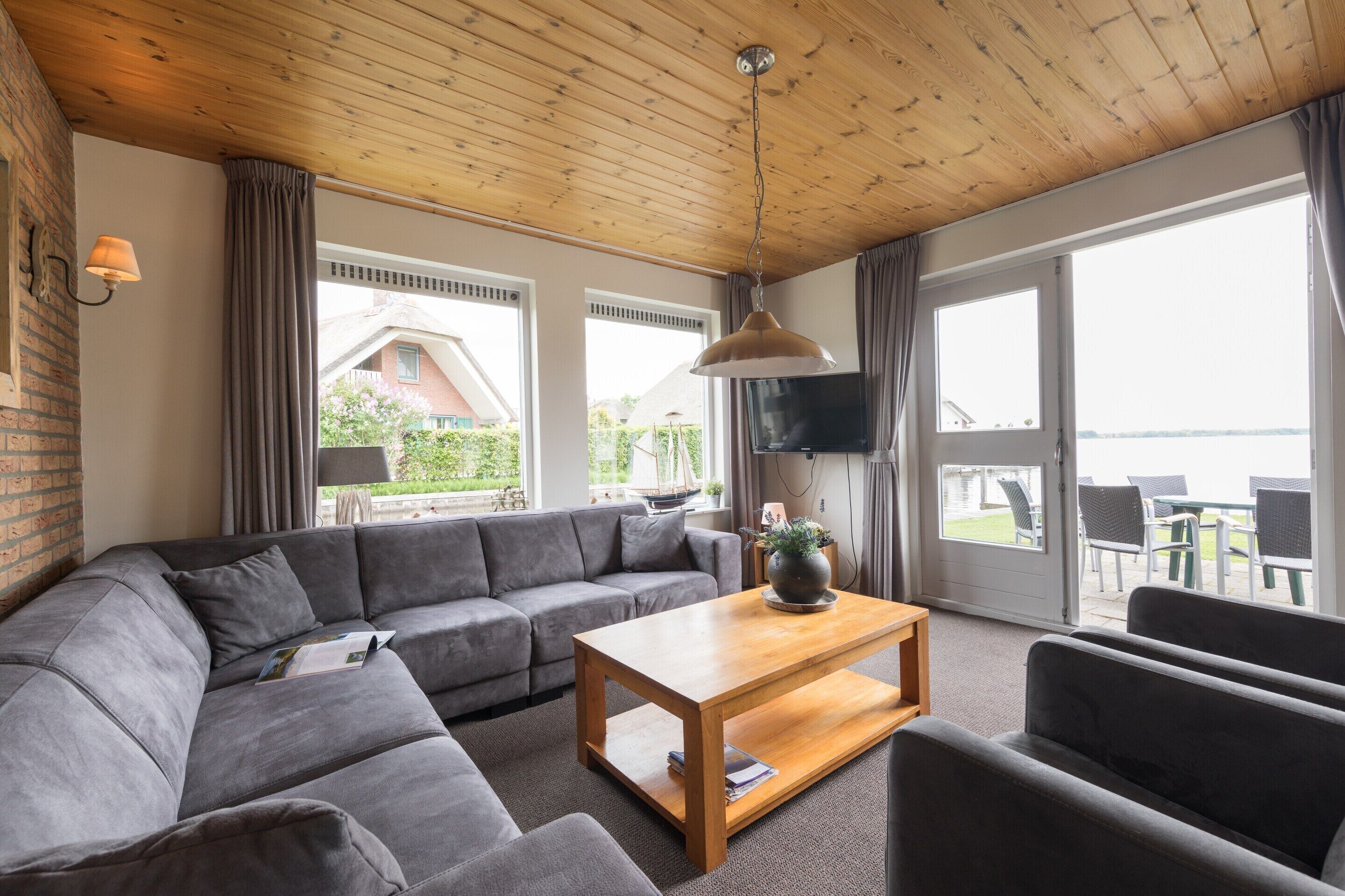 A modern living room with a wooden ceiling, grey sofa, wooden coffee table, and large windows offering a view of the outdoors.