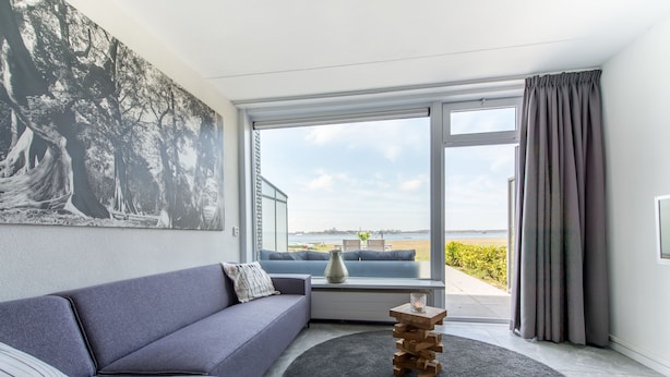Ein stilvolles Wohnzimmer mit grauem Sofa, einem großen Fenster mit Blick auf den Strand und einem Bild an der Wand.