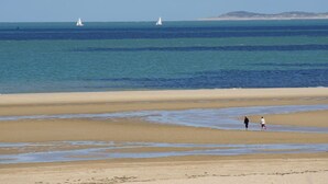 Ein Strand mit zwei Segelbooten in der Ferne und zwei Personen, die am Ufer entlanggehen.