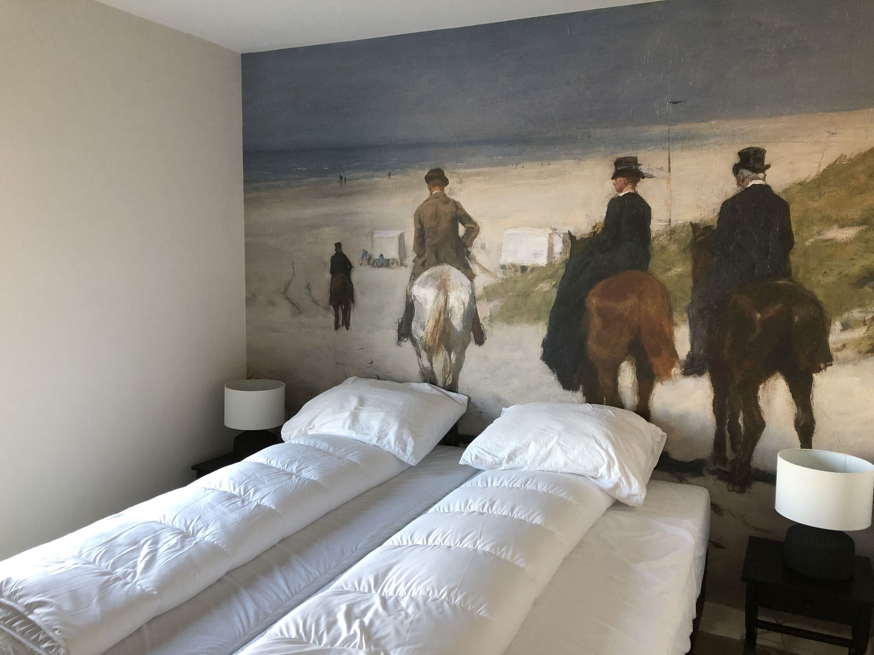 A room with two beds, a large painting of a beach scene, and a lamp on each side of the beds.