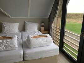 Habitación de hotel con dos camas, una mesita de noche con lámpara y vistas a un paisaje verde a través de una puerta de balcón.