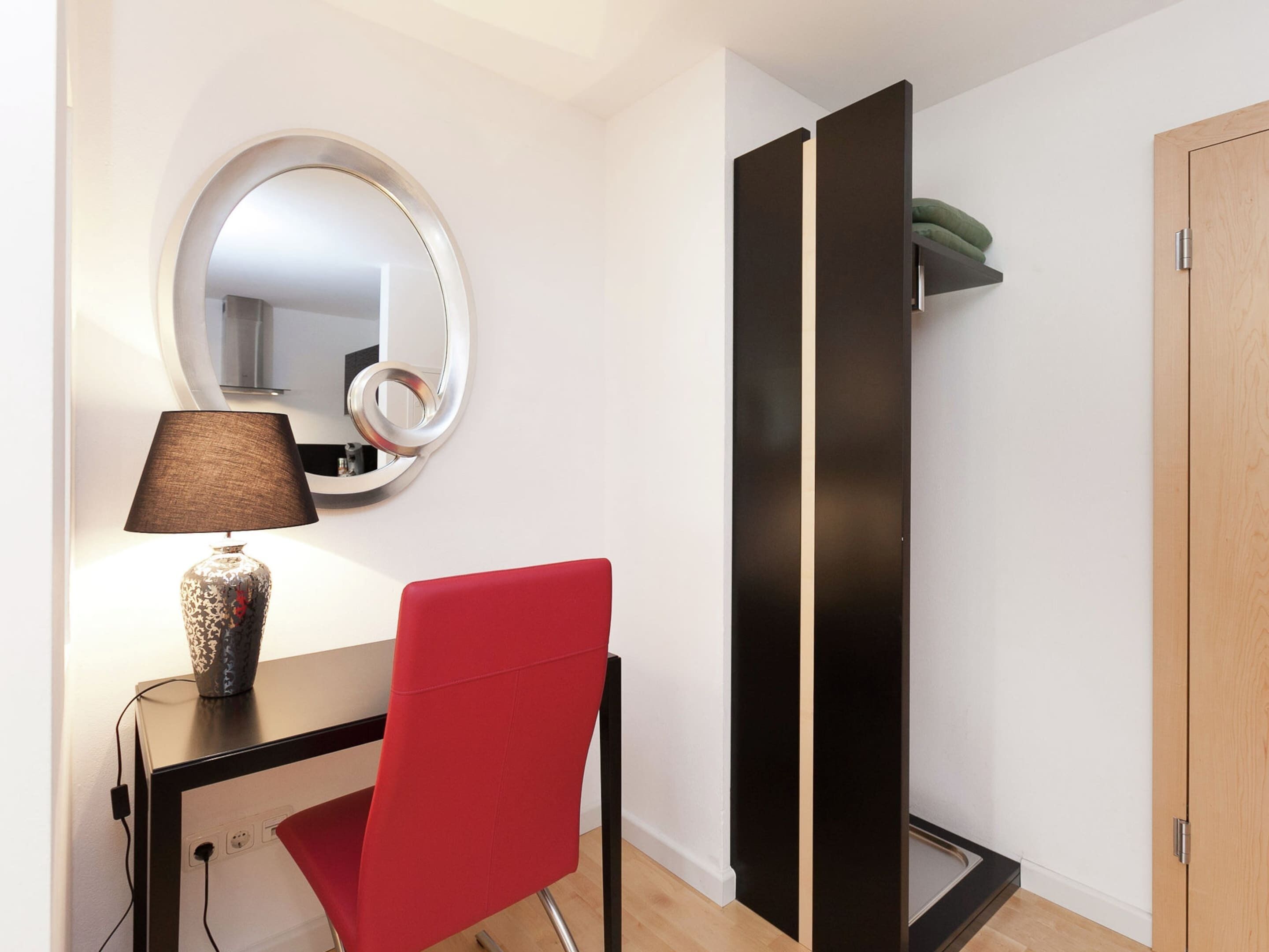 A room with a round mirror, a red chair, a small table with a lamp, and a black wardrobe.