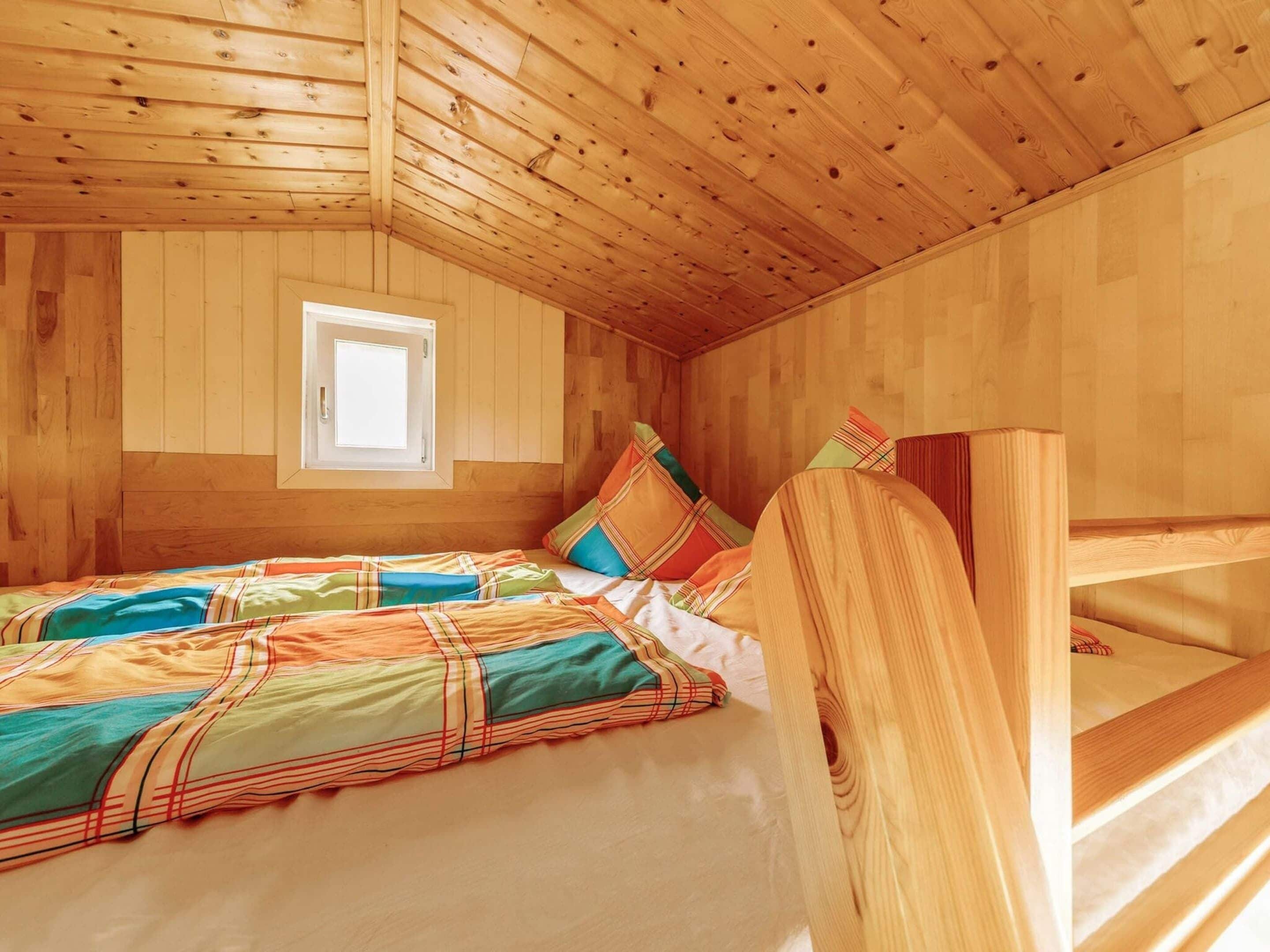 A wooden cabin room with a bed, colorful bedding, a window, and a bunk bed.