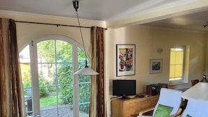 A living room with a wooden table, a sofa, and two chairs with green cushions. There is a TV on a wooden cabinet, a painting on the wall, and a window with curtains. - Tidy House With Sauna and Steam Bath, in a Forest (Brandscheid)