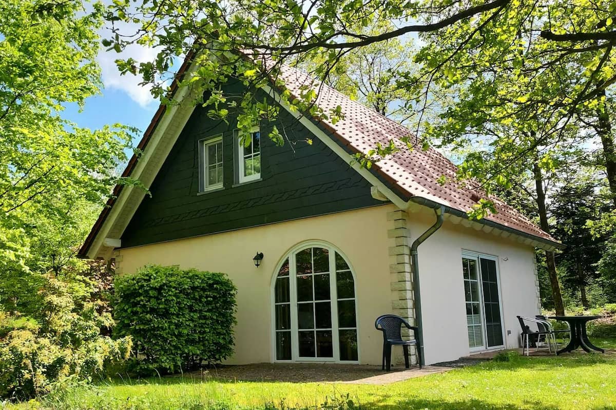 A two-story house with a green roof and beige walls, surrounded by lush greenery.