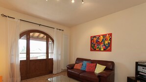 Ein Wohnzimmer mit einem braunen Ledersofa, bunten Kissen, einem Glastisch, einem großen Fenster mit Vorhängen, einer Heizung und einem farbenfrohen abstrakten Gemälde an der Wand.
