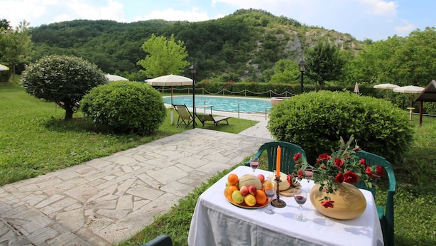 A poolside table with fruit and candles, surrounded by greenery and a view of a pool and hills. - Apartment in Marche With Pool and Privacy (Apecchio)