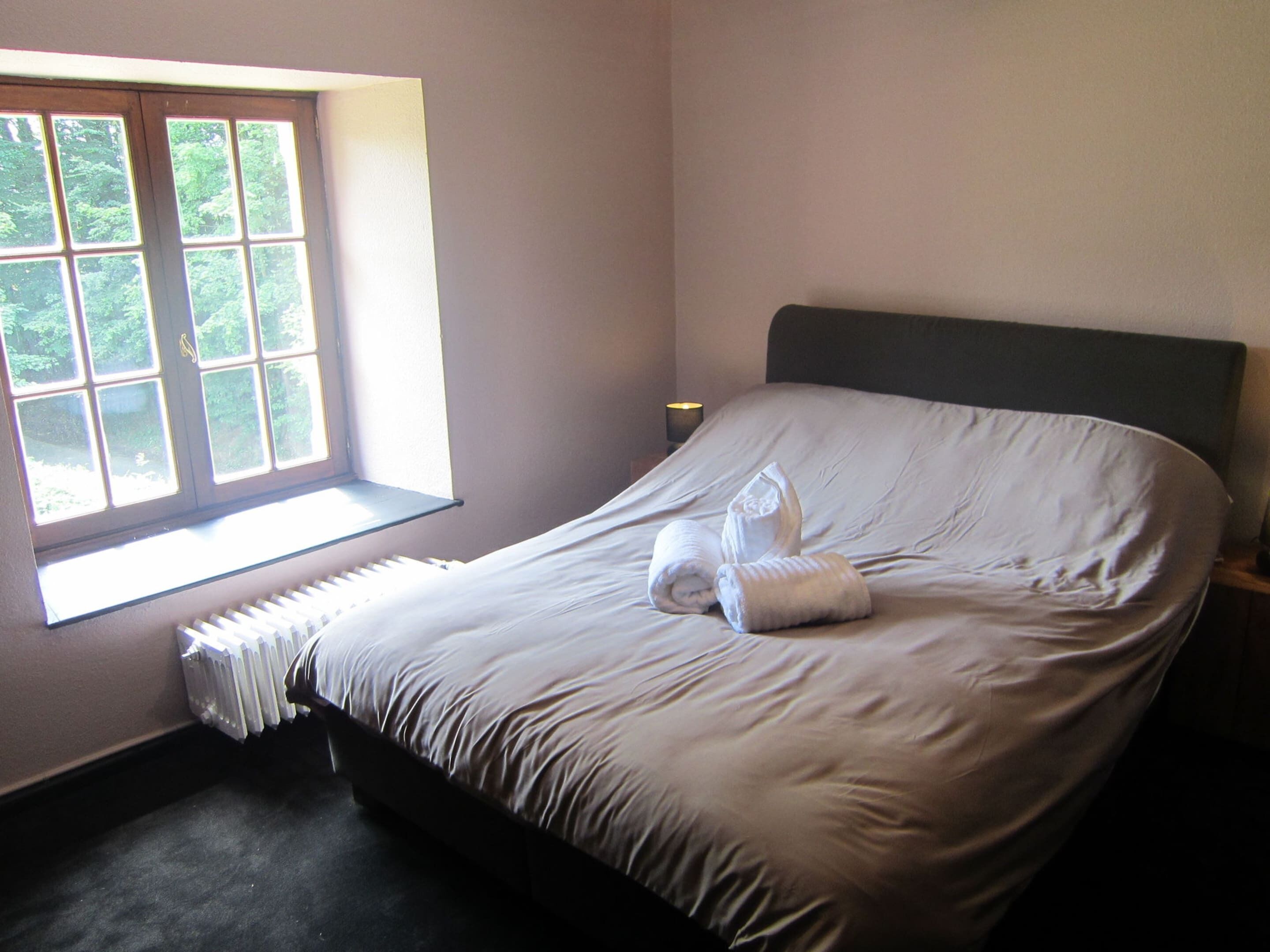 Una cama bien hecha con una toalla enrollada, una ventana que ofrece vistas a zonas verdes y un radiador debajo de la ventana.