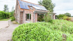 Una casa in mattoni con tetto spiovente ricoperto di pannelli solari, un camino a vista e un giardino con fitta vegetazione.
