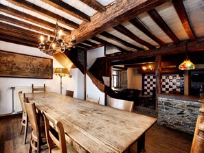 A rustic dining area with a wooden table and chairs, exposed wooden beams, a chandelier, and a stone wall in the background.