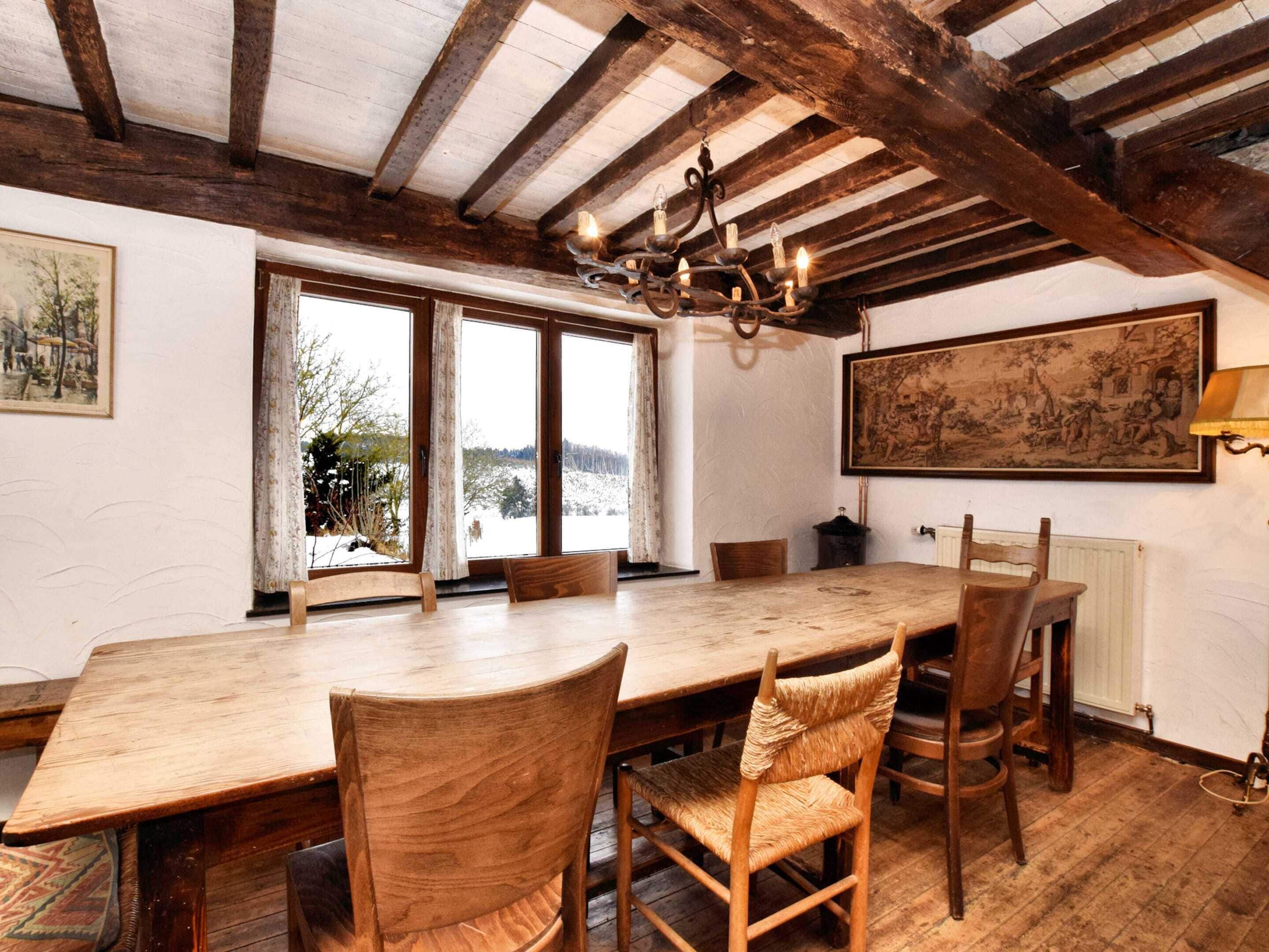 A dining room with a large wooden table, four chairs, a chandelier, and a view of a snowy landscape through large windows.