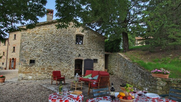 A stone building with a dining area set up outside. There are tables with red and white tablecloths, red chairs, and a wicker chair. A basket of fruit and a bottle of wine are on the table. - Quaint Cottage With Pool (Anghiari)
