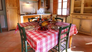 A dining area with a checkered tablecloth, wooden chairs, and a variety of fruits and bottles on the table. - Belvilla by OYO La Verbena (Montalcino)