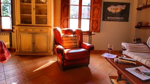 A cozy living room with a leather armchair, a wooden cabinet, a sofa, and a table with books and a wine glass. - Belvilla by OYO La Verbena (Montalcino)