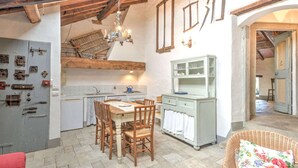 A rustic kitchen with exposed wooden beams, a dining table with chairs, and a wicker chair with a floral cushion.