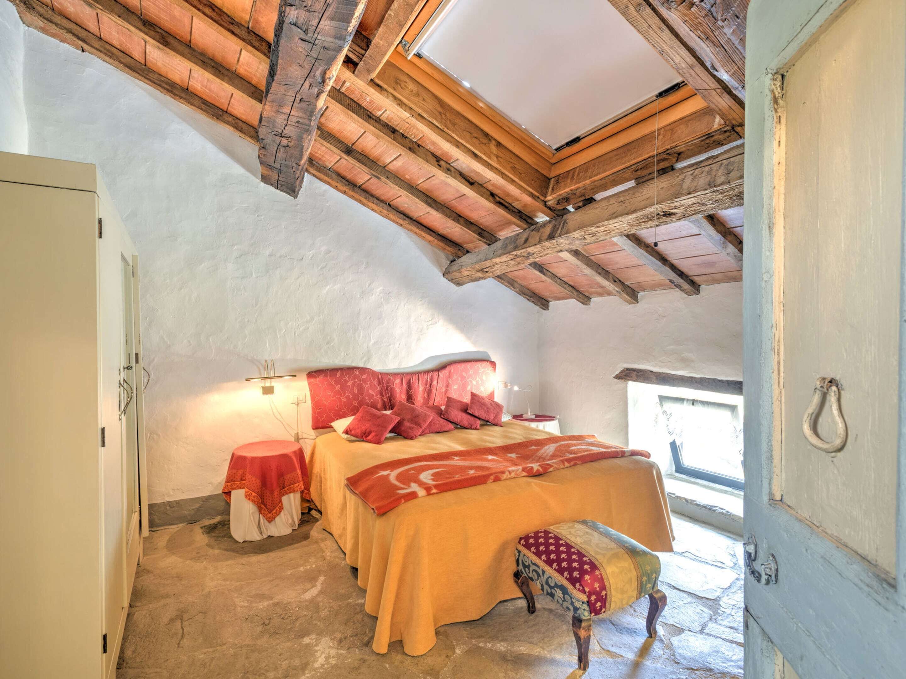 A bedroom with a sloped ceiling, a bed with red bedding, a small table with a red tablecloth, and a bench with patterned upholstery.