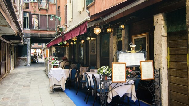 An outdoor café with tables and chairs, a menu board, and a blue carpet. - Belvilla by OYO Paradiso (Venice)