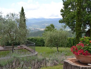 A garden with a circular stone bench, a large potted plant with red flowers, and a view of distant mountains and a town. - Authentic Farmhouse with Pool, Hot Tub, Playground, Garden (Poppi)