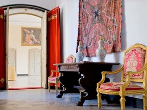A room with a red curtain, a table with vases, a chair, and a painting on the wall.